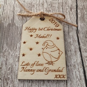 May include: A wooden Christmas ornament with a cute cartoon bird wearing a Santa hat. The ornament is personalized with the text "Happy 1st Christmas Mabel!! Lots of love Nanny and Grandad XXX" and the year "2019".