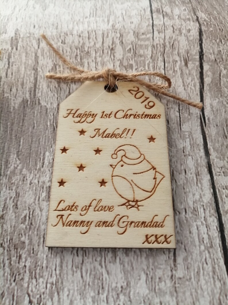 May include: A wooden Christmas ornament with a cute cartoon bird wearing a Santa hat. The ornament is personalized with the text "Happy 1st Christmas Mabel!! Lots of love Nanny and Grandad XXX" and the year "2019".