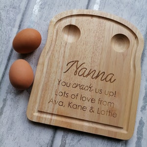 May include: A wooden egg tray shaped like a slice of toast with two egg-shaped indentations. The tray is engraved with the text "Nanna You crack us up! Lots of love from Ava, Kane & Lottie".