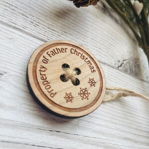 May include: A wooden ornament shaped like a button with the text "Property of Father Christmas" and snowflakes around the edge.