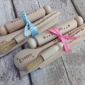 May include: Two wooden baking sets, each with a rolling pin, spoon, and brush, tied with ribbons. One set has a blue ribbon, the other a pink ribbon. The rolling pins are engraved with stars and the names "Ethan" and "Evelyn".