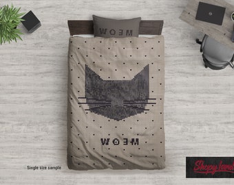 May include: A beige knitted throw blanket with a black cat face design and the word "Meow" written in black. The blanket is on a bed with a pillow that also says "Meow".
