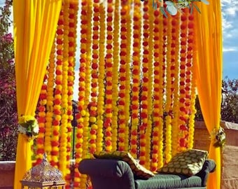 May include: A wedding ceremony backdrop decorated with yellow and orange marigold flower garlands, a green velvet couch, and a yellow and red rug.