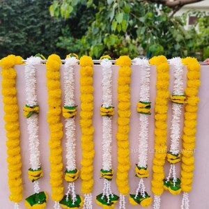May include: A display of decorative garlands. The garlands feature alternating strands of yellow and white flowers, with green and yellow accents. The garlands are hanging against a light pink wall, with greenery in the background.