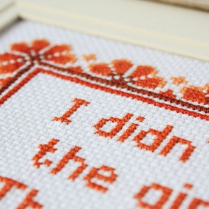 May include: Cross-stitch artwork in a white frame. The piece features orange text that reads "I didn't the girl". The border is decorated with an orange and brown floral design. The background is white fabric.