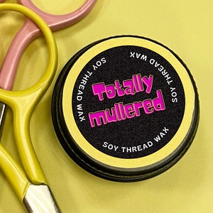 May include: A round container of soy thread wax with a black lid and a gold label. The label says "Soy Thread Wax" and "Totally Mullered" in pink letters.