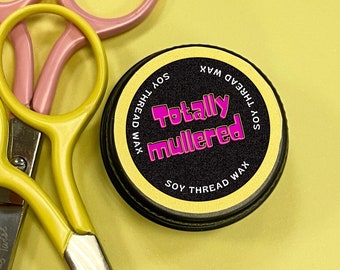 May include: A round container of soy thread wax with a black lid and a gold label. The label says "Soy Thread Wax" and "Totally Mullered" in pink letters.