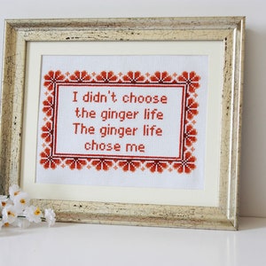 May include: Framed cross-stitch artwork with the text "I didn't choose the ginger life. The ginger life chose me." The text is in red, surrounded by a red floral border, and framed in a gold-toned frame.