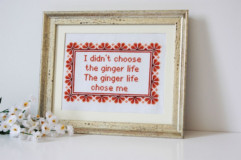 May include: Framed cross-stitch artwork with the text "I didn't choose the ginger life. The ginger life chose me." The text is in red, surrounded by a red floral border, and framed in a gold-toned frame.
