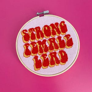 May include: A pink and white embroidery hoop with the text "Strong Female Lead" in red and yellow lettering.
