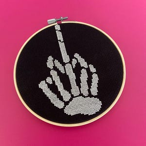 May include: A black and white cross-stitch embroidery of a skeleton hand giving the middle finger, framed in a wooden hoop.