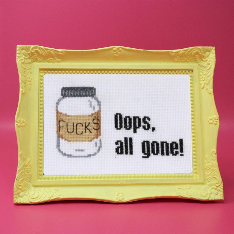 May include: Cross-stitch art in a yellow frame. The artwork features a jar with the word "FUCKS" on it, and the text "Oops, all gone!" on a white background. The frame is a light yellow color.