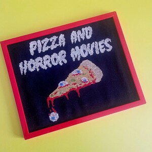 Może przedstawiać: Czarny ramka z czerwonym obramowaniem wokół haftu krzyżykowego. Haft przedstawia tekst "Pizza and Horror Movies" i kawałek pizzy z oczami i kapiącym czerwonym sosem.