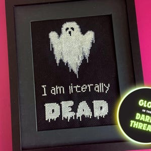 May include: A black framed cross-stitch with a white ghost and the text "I am literally DEAD". The frame is on a pink background. A glow-in-the-dark thread label is in the lower right corner.