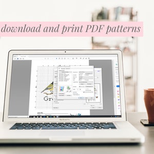 May include: A laptop displays a cross-stitch pattern of a bird, with a software window open. The text "Easily download and print PDF patterns" is above the laptop. A brown mug sits on a coaster to the right.
