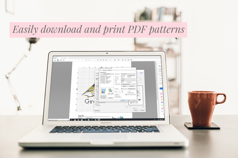 May include: A laptop displays a cross-stitch pattern of a bird, with a software window open. The text "Easily download and print PDF patterns" is above the laptop. A brown mug sits on a coaster to the right.