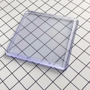 Könnte beinhalten: Ein klarer, quadratischer Untersetzer mit abgerundeten Ecken. Der Untersetzer besteht aus transparentem Kunststoff und hat einen leicht erhöhten Rand. Der Hintergrund ist eine weiße Oberfläche mit einem schwarzen Gittermuster.