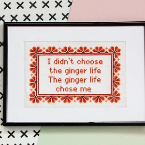 May include: Framed cross-stitch art with the text "I didn't choose the ginger life. The ginger life chose me." The text is in red, framed by a red floral border, and set against a white background. The frame is black.