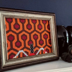 Può includere: Un ricamo a punto croce incorniciato che rappresenta l'iconico motivo del tappeto dell'"Overlook Hotel" del film "Shining". Il tappeto è arancione e rosso con una silhouette nera di una figura in piedi al centro.