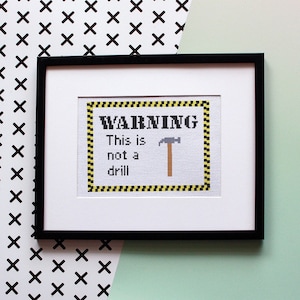 Puede incluir: Un diseño de punto de cruz enmarcado con una señal de advertencia amarilla y negra que dice "WARNING This is not a drill" con una imagen de martillo de dibujos animados.