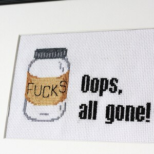 May include: Cross-stitch art framed in black. The artwork features a jar with the word "FUCKS" on the label, and the text "Oops, all gone!" in black. The jar is white with a brown label and a gray lid.