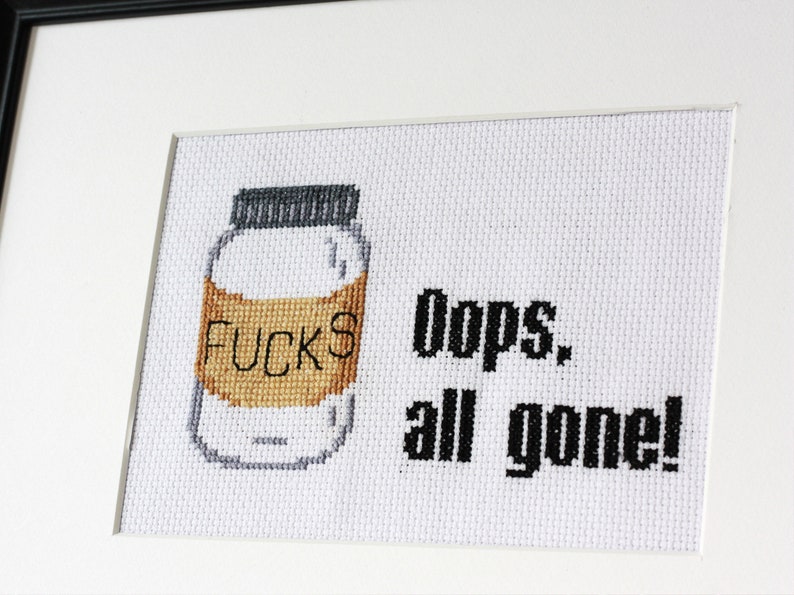 May include: Cross-stitch art framed in black. The artwork features a jar with the word "FUCKS" on the label, and the text "Oops, all gone!" in black. The jar is white with a brown label and a gray lid.