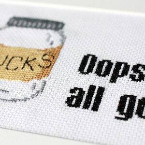 May include: Cross-stitch art on white fabric. A jar with the word "FUCKS" stitched on it is on the left. The text "Oops. all gone" is stitched in black on the right. The jar has a gray lid and a tan filling.