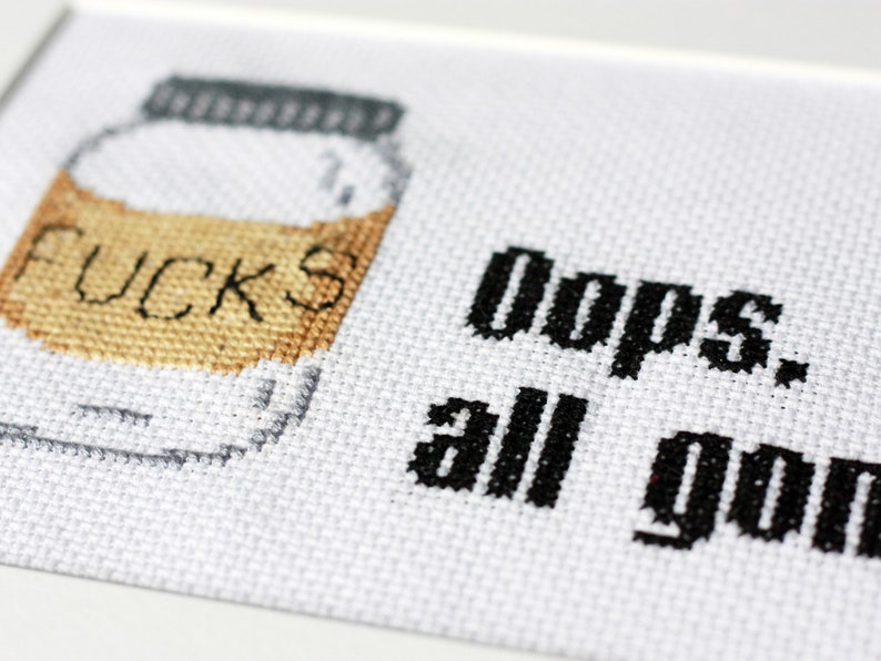 May include: Cross-stitch art on white fabric. A jar with the word "FUCKS" stitched on it is on the left. The text "Oops. all gone" is stitched in black on the right. The jar has a gray lid and a tan filling.