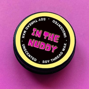 May include: A black and yellow round container with the text "IN THE NUDDY" in pink letters. The container is labelled "Unscented Soy Thread Wax".