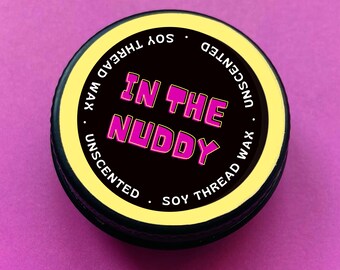May include: A black and yellow round container with the text "IN THE NUDDY" in pink letters. The container is labeled "Unscented Soy Thread Wax".