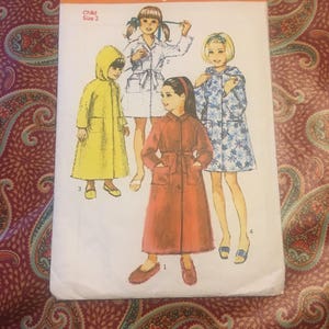 May include: A vintage Simplicity sewing pattern for children's robes. The pattern number is 8434 and the price is 25p. The pattern includes four different styles of robes, including a hooded robe, a belted robe, and a robe with a floral print. The pattern is for child size 2.
