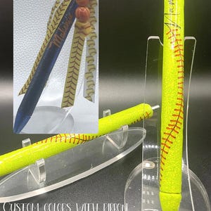 May include: A yellow pen with red stitching, resembling a softball, is displayed on a clear stand. The pen has a glittery texture and is accompanied by a pen with a blue and yellow ribbon and beads. The text on the pen says "Melanie".