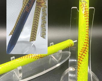 May include: A yellow pen with red stitching, resembling a softball, is displayed on a clear stand. The pen has a glittery texture and is accompanied by a pen with a blue and yellow ribbon and beads. The text on the pen says "Melanie".