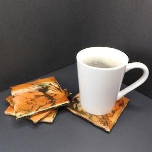 May include: Set of four orange and black abstract coasters with a white coffee mug on top of one coaster.