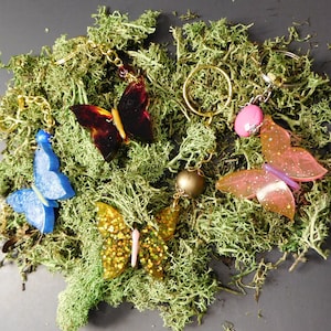 May include: A collection of butterfly-shaped keychains in various colors, including blue, gold glitter, and translucent pink. Each keychain features a gold-colored chain and is set against a backdrop of green moss.