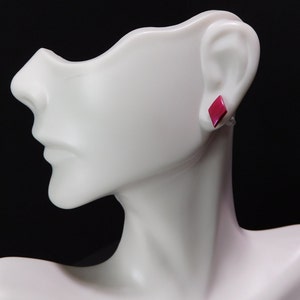 May include: A pair of bright pink, diamond-shaped stud earrings. The earrings are displayed on a white mannequin head against a black background. The earrings have a glossy finish and are a vibrant shade of pink.