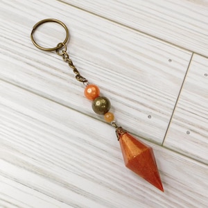 May include: A gold-toned key chain with a sparkly orange and gold geometric charm, three orange beads, and a gold-toned ring.