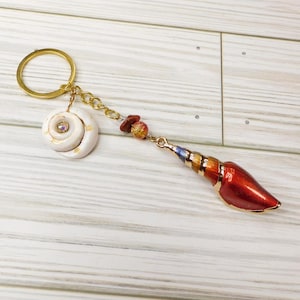 May include: A gold key chain with a white seashell charm and a red, gold, and blue seashell charm. The key chain is on a white wooden surface.