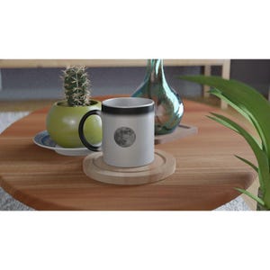 May include: A white ceramic mug with a black handle and rim, featuring a detailed image of the moon. The mug sits on a wooden coaster on a wooden table, with a cactus in a green pot and a decorative vase in the background.