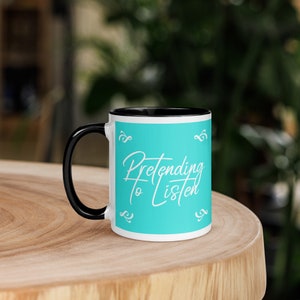 Puede incluir: Una taza de café blanca con un borde y un asa negros. La taza tiene un fondo azul turquesa con el texto "Pretending to Listen" en escritura cursiva blanca.