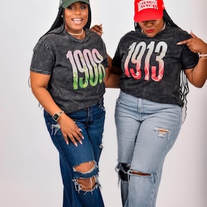 May include: Two charcoal acid-wash t-shirts featuring large graphic numbers. One shirt displays 1908 in pink and green, paired with a green baseball cap. The other shirt displays 1913 in red and white, paired with a red baseball cap and distressed jeans.