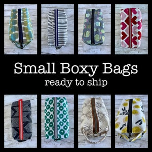 May include: Collection of small boxy bags in various patterns, including geometric shapes, stripes, and floral designs. Each bag features a zipper closure and is ready to ship. The bags are displayed on a light wood-grain surface.