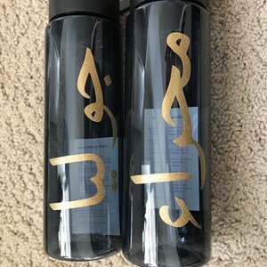 May include: Two black water bottles with gold lettering. The lettering on the left bottle says "B" and the lettering on the right bottle says "Brooke".