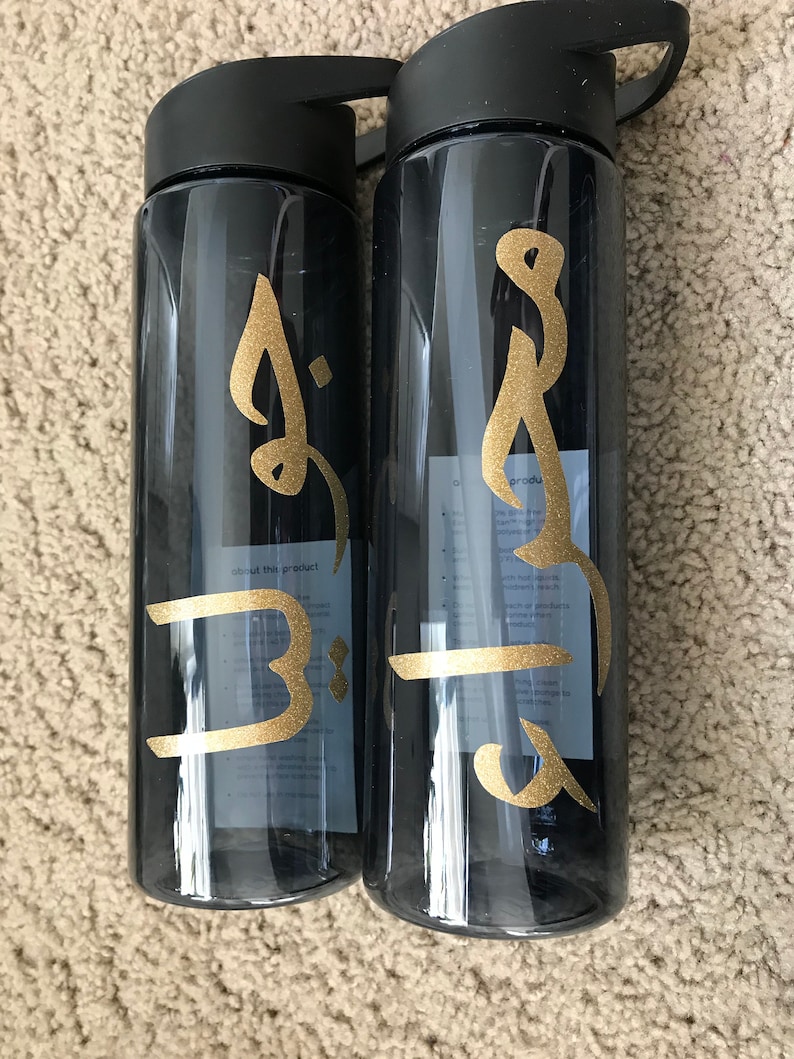 May include: Two black water bottles with gold lettering. The lettering on the left bottle says "B" and the lettering on the right bottle says "Brooke".