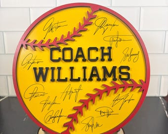 May include: A round, yellow softball-themed sign with red stitching and border. The sign features the text "COACH WILLIAMS" in black, surrounded by signatures. It is displayed on a wooden stand.