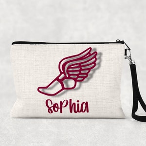 May include: A cream-colored zippered pouch with a black zipper and wrist strap. The pouch features a maroon graphic of a winged foot and the name "Sophia" in a matching maroon font. The pouch is made of a textured fabric.