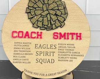 May include: A round wooden plaque with the words "COACH SMITH" in pink letters. The plaque features a gold glitter pom-pom design and the words "EAGLES SPIRIT SQUAD" with a list of names. The text "THANK YOU FOR A GREAT SEASON" is at the bottom.