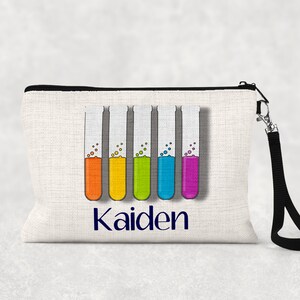 以下が含まれることがあります： 黒いジッパーと黒いストラップが付いた白いキャンバス製のジッパーポーチ。ポーチには、異なる色の液体で満たされた6つの試験管のカラフルなグラフィックが描かれています。テキスト「Kaiden」は、グラフィックの下に青色で印刷されています。