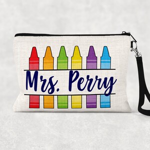 Puede incluir: Una bolsa de lona blanca con cremallera negra y correa negra. La bolsa tiene un diseño de una fila de lápices de colores con el texto "Mrs. Perry" en azul.