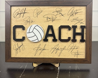 May include: Wooden framed sign with the word "COACH" in black letters and a white and black volleyball. The sign has a light wood background with signatures and is supported by a wooden stand.